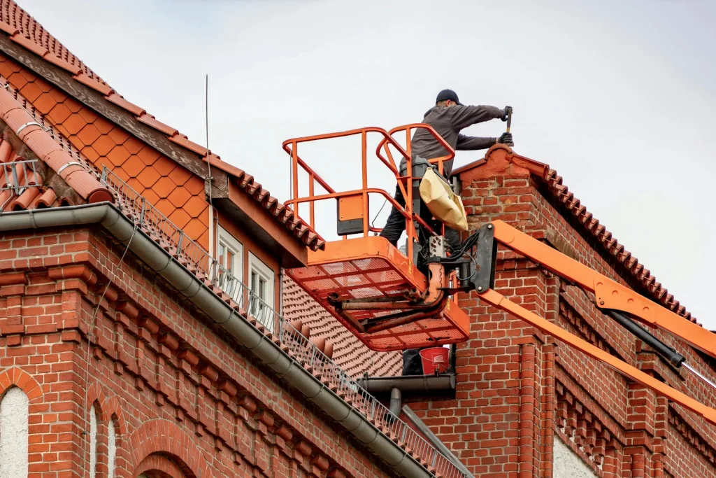 Professional contractor performing roof leak repair on a tile roof near a chimney using elevated access equipment.